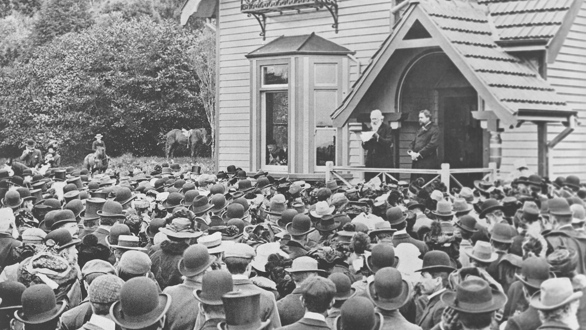 Sir John Logan Campbell addressing the crowd at the official opening of Cornwall Park in 1903, with him on the porch of Huia Lodge is the Honourable E Mitchelson, Mayor of Auckland. Auckland Libraries Heritage Collections 7-A253